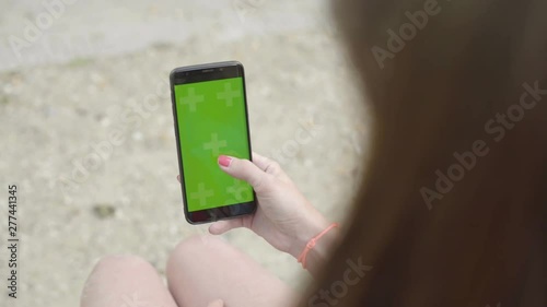 Woman using smartphone with Green Screen Sitting on bench in park, Swiping Scrolling on Touch screen with Tracking Markers. Browsing Internet, Watching Media Content, Videos, Blogs, Social Networks.