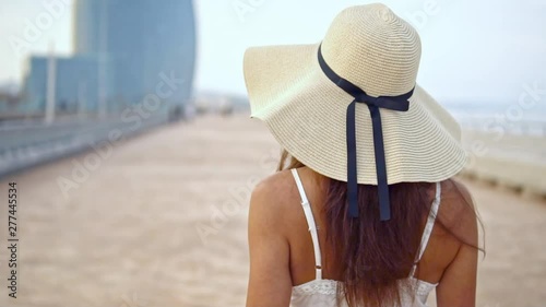 Pretty woman wearing white dress and pamela hat walking over the beach terrace at sea. Happy summer vacation photo