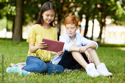 Smiling schoolgirl in yellow T-shirt sitting on grass and showing her project on tablet to her classmate