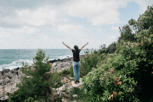 A girl stands on a stone seashore, admires the sea and raises her hands up. She feels free and happy.