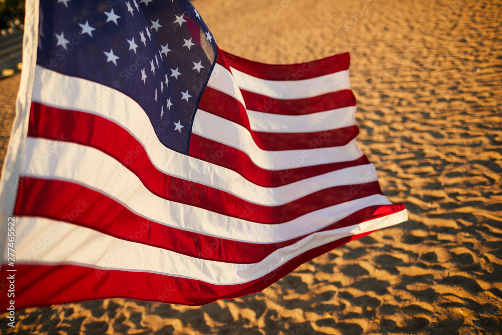 Obraz premium Close-up of American flag with stripes and stars blowing in wind on summer beach at sunset