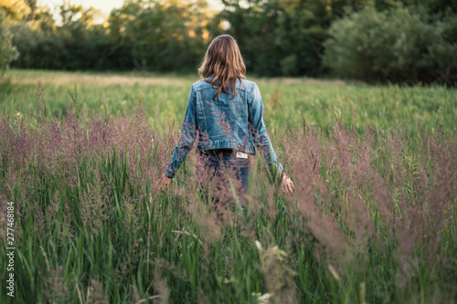 Caucasian girl with long brown hair wearing a denim jacket walking through a field of tall grass in the evening sun
