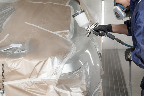 A male worker paints with a spray gun a part of the car body in silver after being damaged at an accident. Rear fender from the vehicle during the repair in workshop. Auto service industry professions