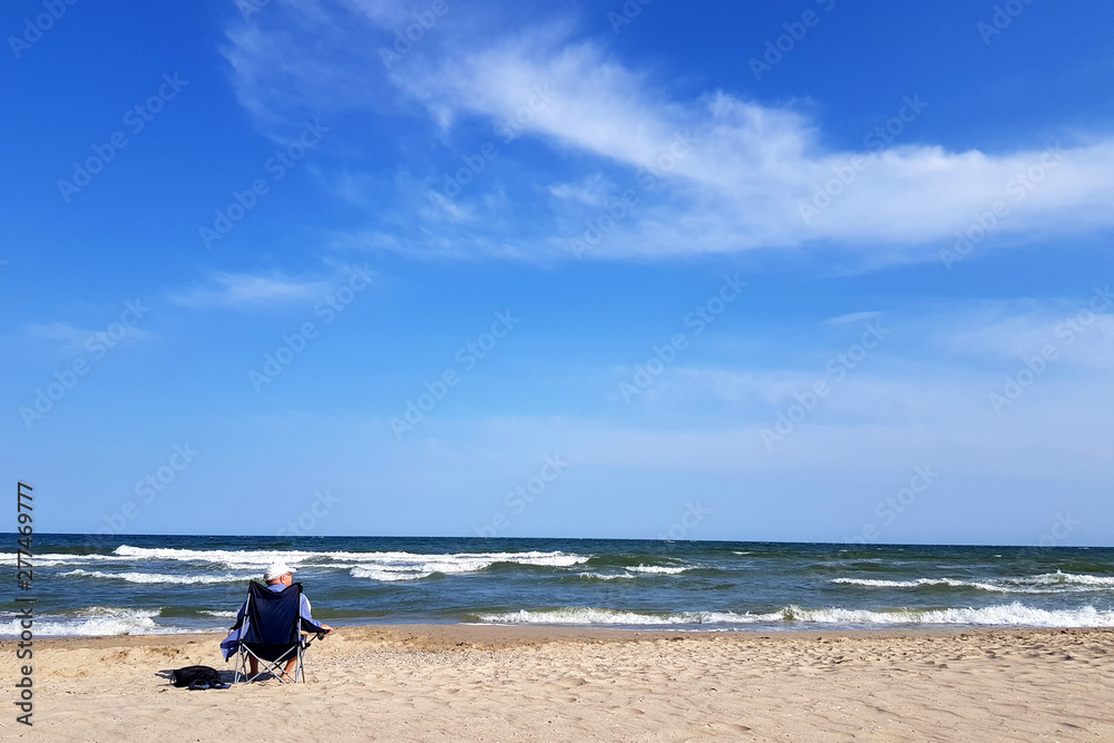 Man with hat sitting on a beach chair and holding a beer bottle, on a beach next to a sea