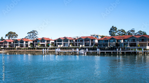 Large waterside houses in suburban community set on riverfront with wooden wharf and boat in foreground, blue sky in background