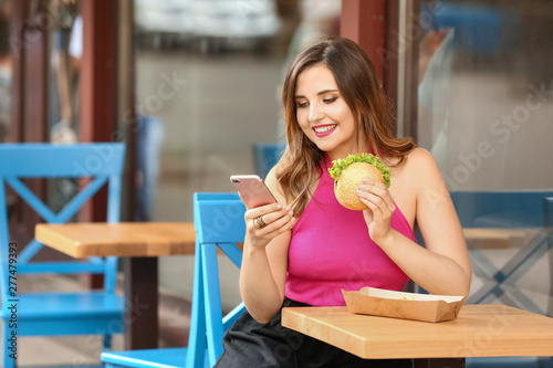 Beautiful young woman with mobile phone eating tasty burger in outdoor cafe