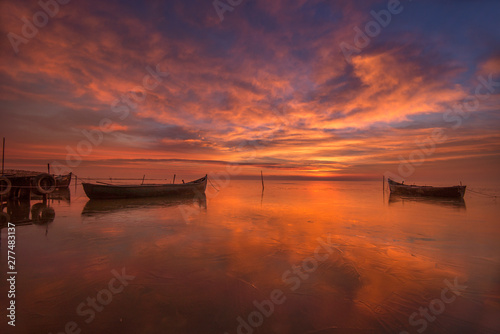 Colorful sunrise at the sea with boats floating on calm water and clouds reflecting on the water