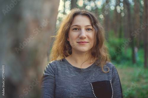 Serious woman looking out from behind tree. Pretty calm woman touching tree and looking at camera in quiet forest