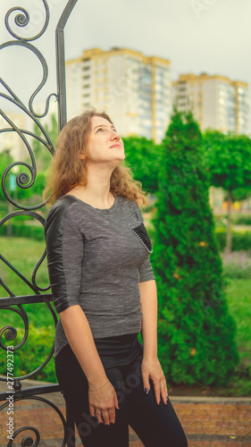 Smiling woman against fence in garden. Content woman looking dreamily away while standing in green park against ornamental iron fence