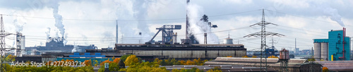 Large panoramic view of an industrial landscape: factory chimneys poring smoke into a cloudy sky while power lines transport electricity. In front the tops of a few autumn colored trees are visible.