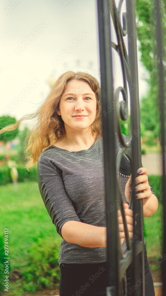 Smiling woman against fence in garden. Content woman looking dreamily standing in green park against ornamental iron fence Smiling woman against fence in garden. Content woman looking dreamily standing in green park against ornamental iron fence