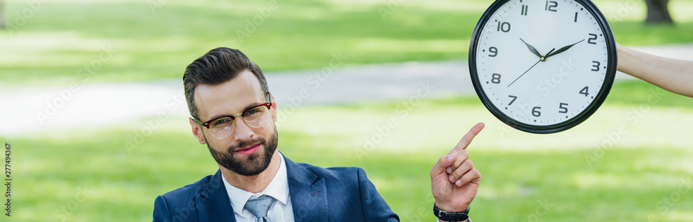 panoramic shot of businessman pointing with finger at clock while sitting in park panoramic shot of businessman pointing with finger at clock while sitting in park