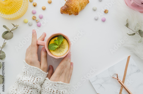 An overhead view of woman holding cup of lemon tea on white table with croissant and candies on white background