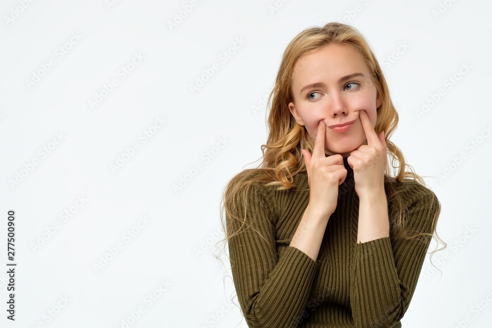 Young upset curly blonde woman making fake smile with her fingers stretching the corners of her mouth. Try to stay positive after failing at something. Young upset curly blonde woman making fake smile with her fingers stretching the corners of her mouth. Try to stay positive after failing at something.