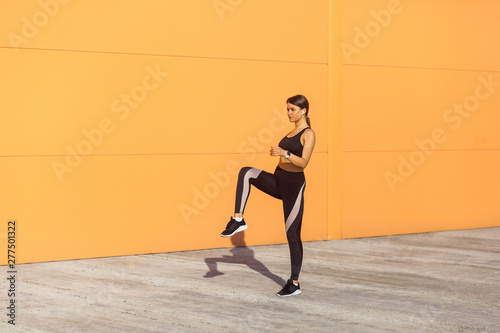 Young attractive sporty woman wearing black sporwear practicing sport exercises in morning on street, standing with raised leg and holding balance, orange wall background, outdoor sport concept