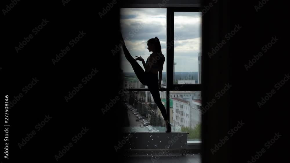 Beautiful scene of stretching sporty young woman - girl doing stretch of her legs, raising one foot above her head against the background of large window in slow motion. Silhouette of girl Beautiful scene of stretching sporty young woman - girl doing stretch of her legs, raising one foot above her head against the background of large window in slow motion. Silhouette of girl