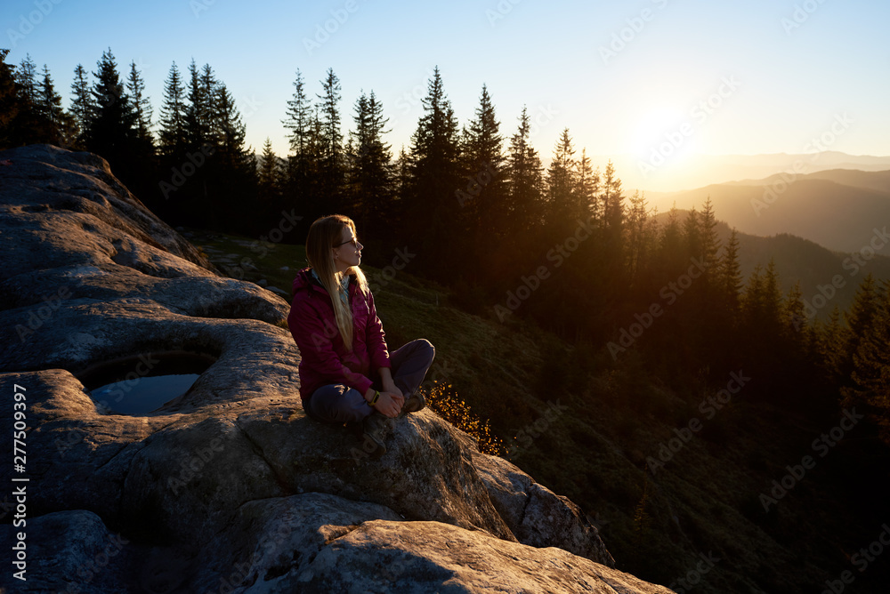 Houng woman traveller sitting on big rock on the top of mountain in the morning. Active female tourist enjoying amazing sunrise. On background forest, rising sun and blue sky Houng woman traveller sitting on big rock on the top of mountain in the morning. Active female tourist enjoying amazing sunrise. On background forest, rising sun and blue sky