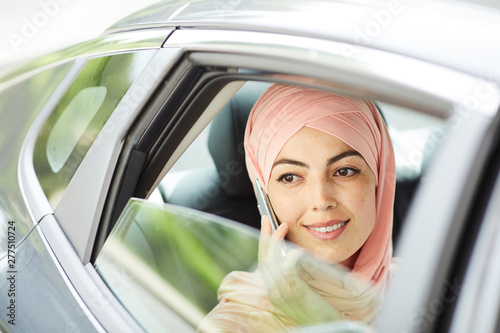 Car window view of positive pensive young Muslim woman in pink hijab talking on phone while riding taxi © Seventyfour