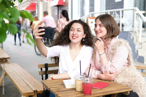 Two young girls take a selfie at a table in a street cafe. Girlfriends communicate and have fun. On the table two summer cocktails in plastic glasses.