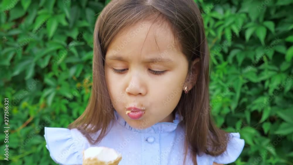 Little cute and funny girl in a blue dress grimaces and eats ice cream. In the background is a green hedge of Virginia creeper (Parthenocissus quinquefolia). Little cute and funny girl in a blue dress grimaces and eats ice cream. In the background is a green hedge of Virginia creeper (Parthenocissus quinquefolia).