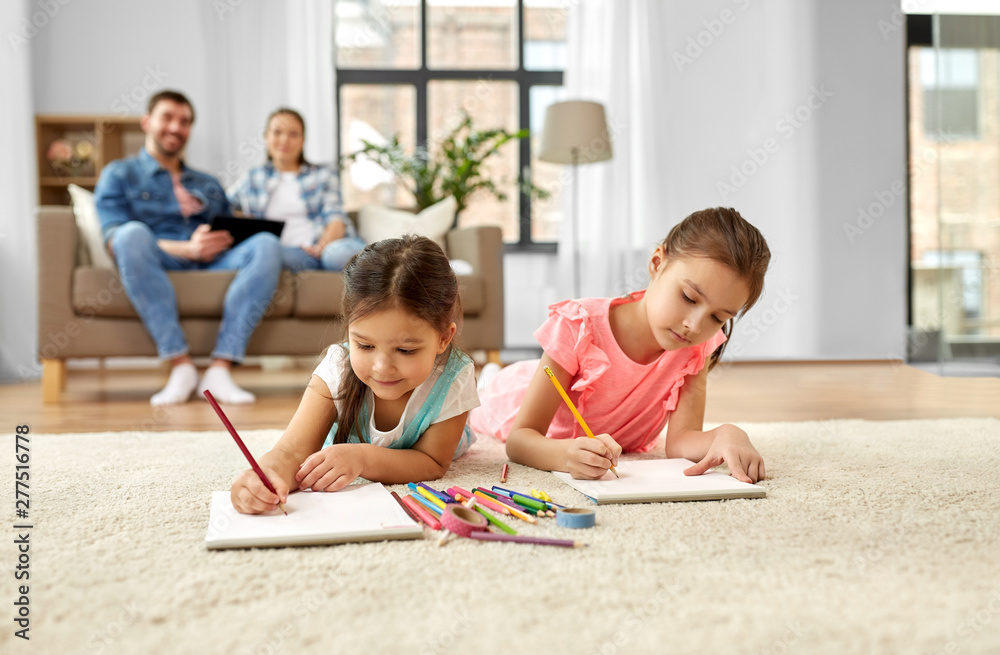 family, leisure and childhood concept - happy sisters lying on floor and drawing in sketchbooks with crayons at home family, leisure and childhood concept - happy sisters lying on floor and drawing in sketchbooks with crayons at home