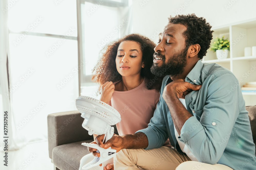 young african man and woman suffering from summer heat and holding blowing electric fan young african man and woman suffering from summer heat and holding blowing electric fan