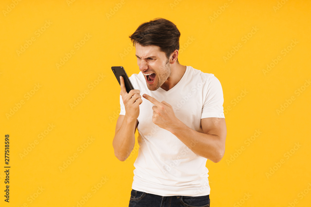 Displeased screaming man in casual white t-shirt talking by mobile phone isolated over yellow background. Displeased screaming man in casual white t-shirt talking by mobile phone isolated over yellow background.