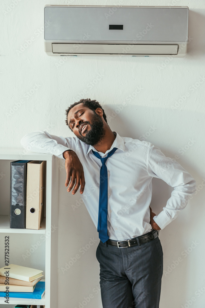 Fototapeta premium exhausted african american businessman standing under conditioner and holding hand on hip