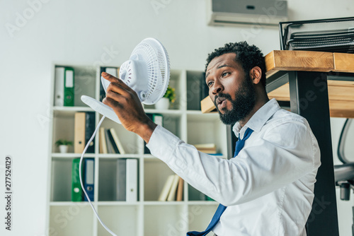 young african american businessman holding blowing electric fan while sitting on floor and suffering from heat in office