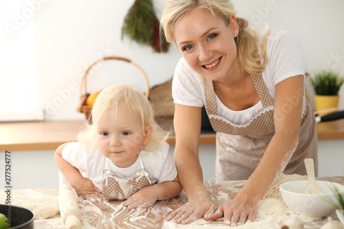 Little girl and her blonde mom in beige aprons playing and laughing while kneading the dough in kitchen. Homemade pastry for bread, pizza or bake cookies. Family fun and cooking concept