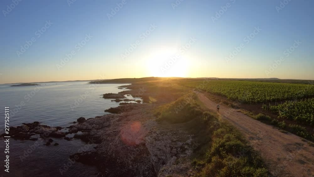 AERIAL Mountain Biker riding his bike along the sea on a dusty road towards the sunset AERIAL Mountain Biker riding his bike along the sea on a dusty road towards the sunset