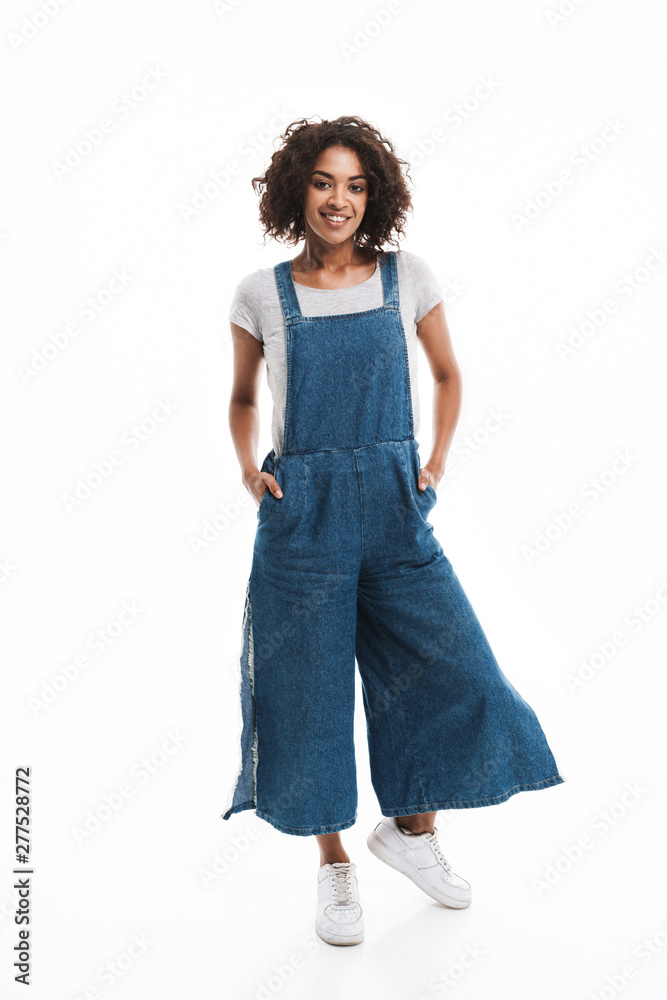 Image of happy african american woman dressed in denim overalls smiling at camera with hands in pockets Image of happy african american woman dressed in denim overalls smiling at camera with hands in pockets