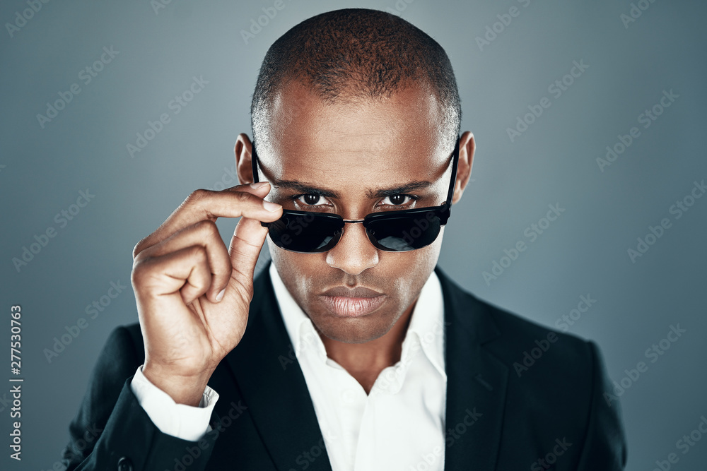 Modern beauty. Charming young African man in full suit looking at camera and adjusting eyewear while standing against grey background Modern beauty. Charming young African man in full suit looking at camera and adjusting eyewear while standing against grey background