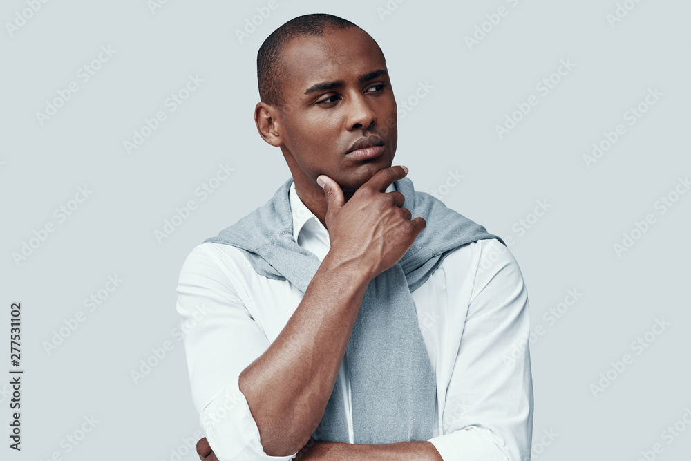 Deep thinking. Handsome young African man looking away and keeping hand on chin while standing against grey background
