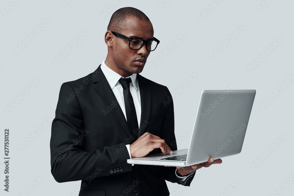 Searching for solution. Young African man in formalwear working using computer while standing against grey background Searching for solution. Young African man in formalwear working using computer while standing against grey background