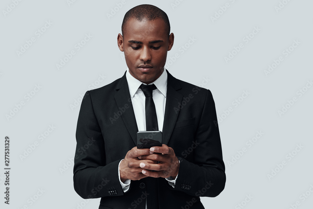 Checking income messages. Charming young African man in formalwear using smart phone while standing against grey background Checking income messages. Charming young African man in formalwear using smart phone while standing against grey background