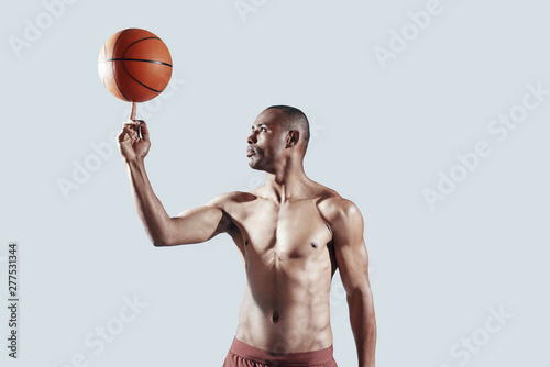 Challenging himself. Handsome shirtless African man spinning ball on finger while standing against grey background