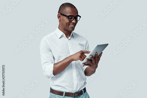  Examining new tablet. Handsome young African man using digital tablet and smiling while standing against grey background
