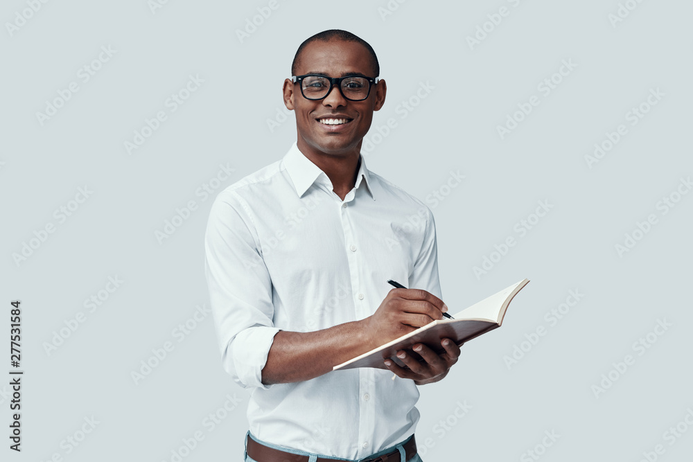 Ready to study. Handsome young African man writing something down while standing against grey background Ready to study. Handsome young African man writing something down while standing against grey background