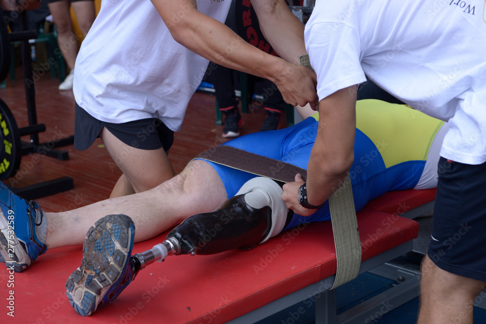 Disabled man with an artificial leg lying on a bench before benching press, assistants fixing his body by belts 