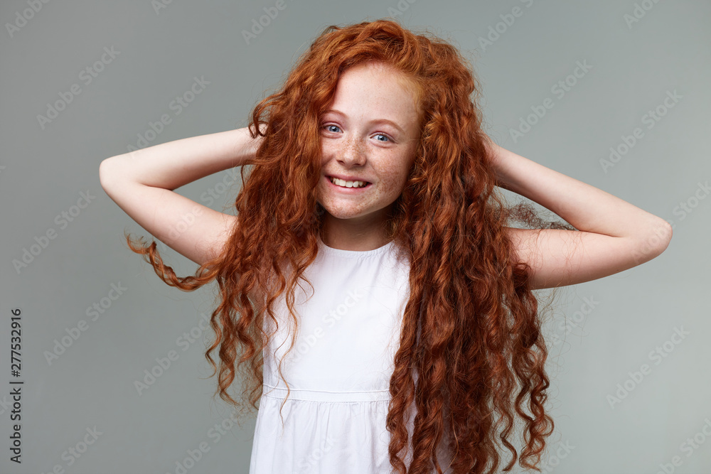 Studio photo of little smiling girl with curly ginger hair and freckles,  stands over gray background and holding hands behind head,straightens thick  hair, looks nice and cute. Stock Photo | Adobe Stock