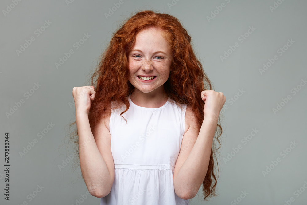Happy little girl with ginger hair and freckles, smiles and looks great, looks at the camera, won the competition, stands over gray background with raised fists up. Happy little girl with ginger hair and freckles, smiles and looks great, looks at the camera, won the competition, stands over gray background with raised fists up.