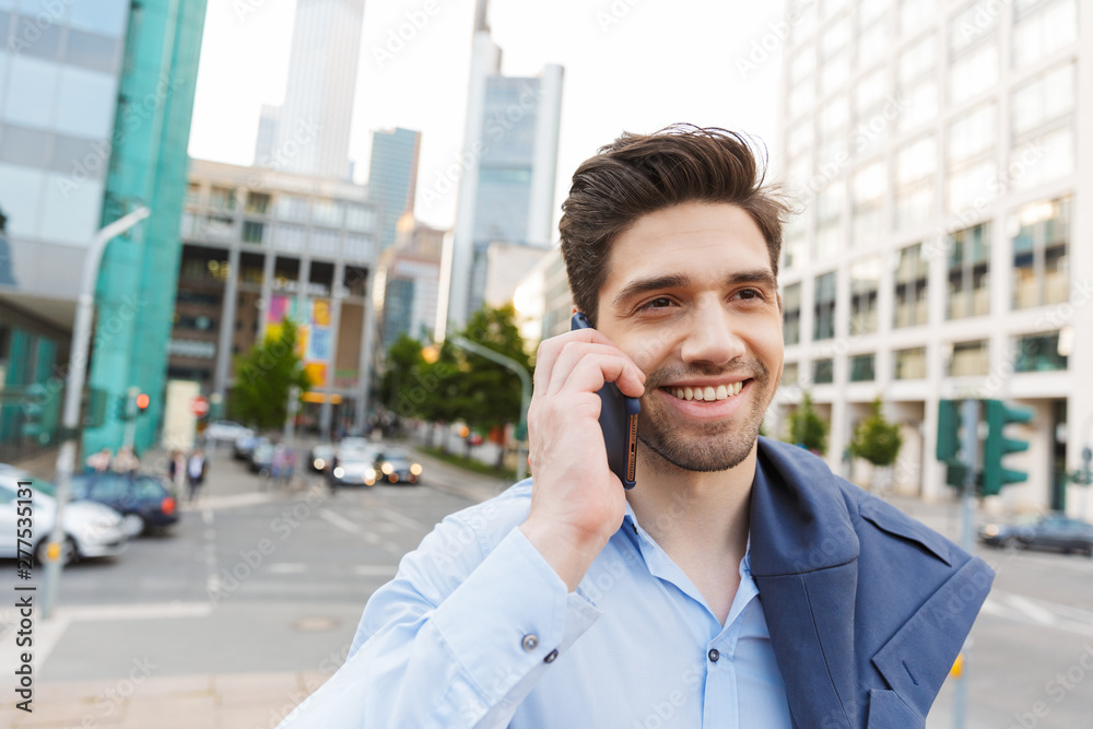 Young positive unshaved businessman walking outdoors on the street talking by mobile phone. Young positive unshaved businessman walking outdoors on the street talking by mobile phone.