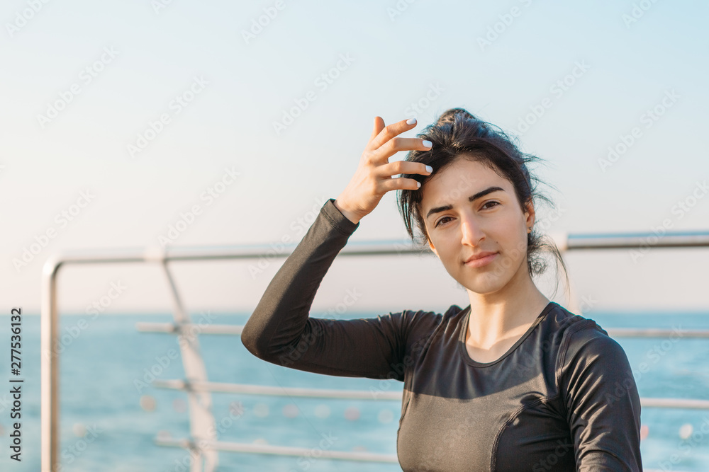 Young beautiful brunette woman in a black sport jumpsuit stands near metal heanrails relaxing after yoga excercising looking at the camera