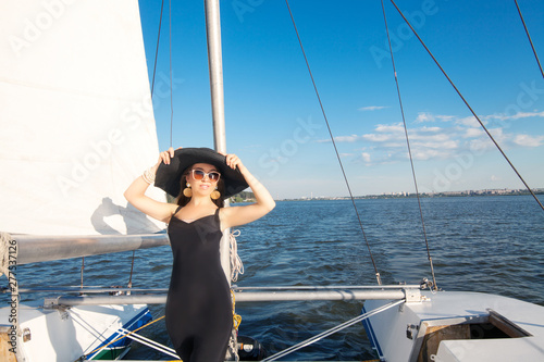  A girl with long hair, a model is standing on a yacht, in a black dress, in a black hat, beside a sail, against a blue sky with space for an inscription. The concept of vacation at sea.
