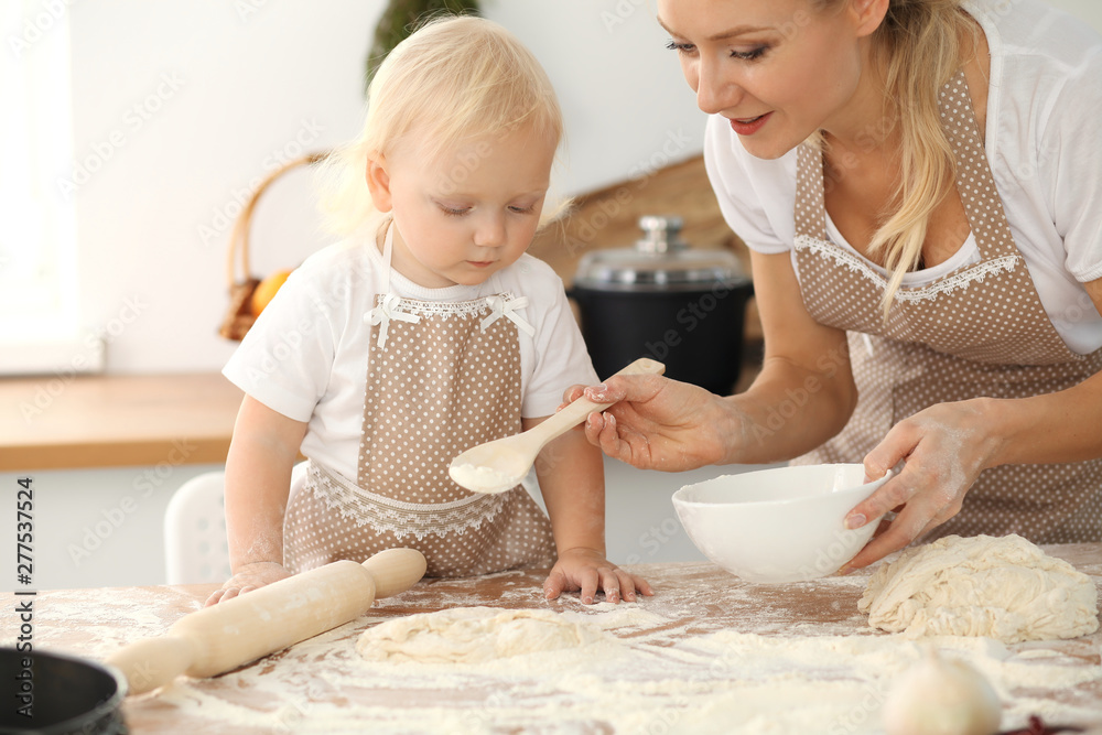 Little girl and her blonde mom in beige aprons playing and laughing while kneading the dough in kitchen. Homemade pastry for bread, pizza or bake cookies. Family fun and cooking concept