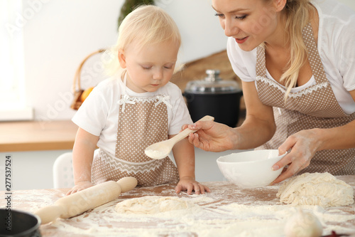 Little girl and her blonde mom in beige aprons playing and laughing while kneading the dough in kitchen. Homemade pastry for bread, pizza or bake cookies. Family fun and cooking concept © rogerphoto