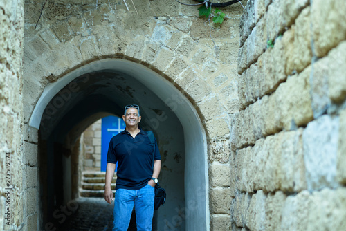 Smiling trendy tourist senior with blue jeans and sunglasses standing by old ancient city street with brick historic stone wall.