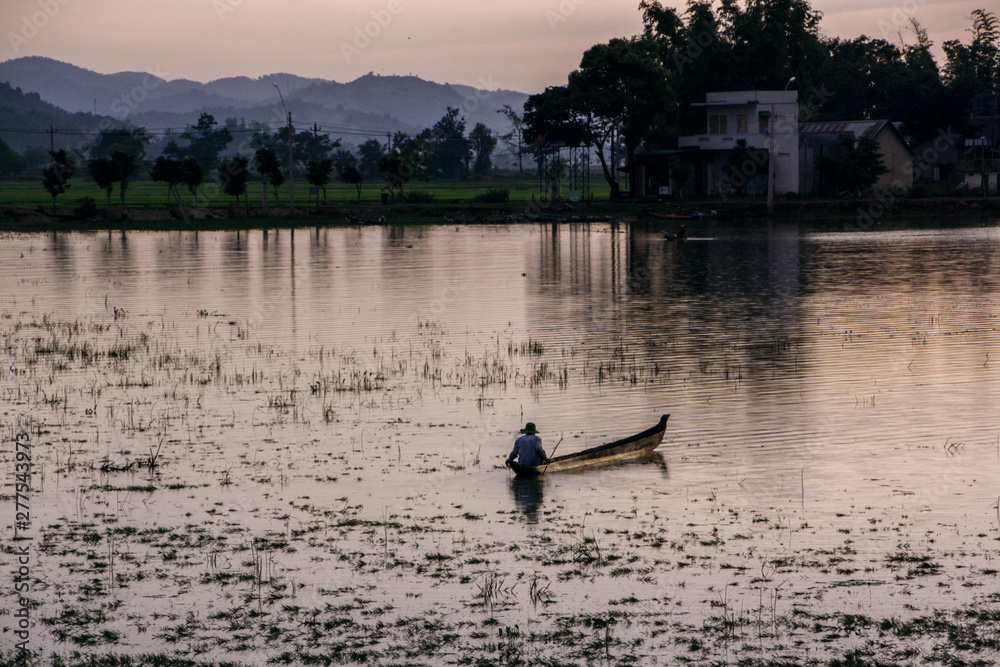 Naklejka premium Vietnamese fisherman sails on a low boat along the shore past the houses. Soon the sunset will begin, twilight lighting