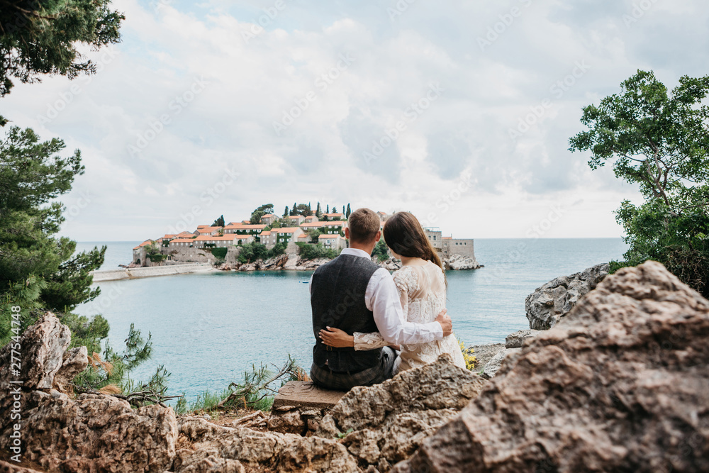 A man and a woman admire a beautiful view of the island of Sveti Stefan in Montenegro. He hugs her.